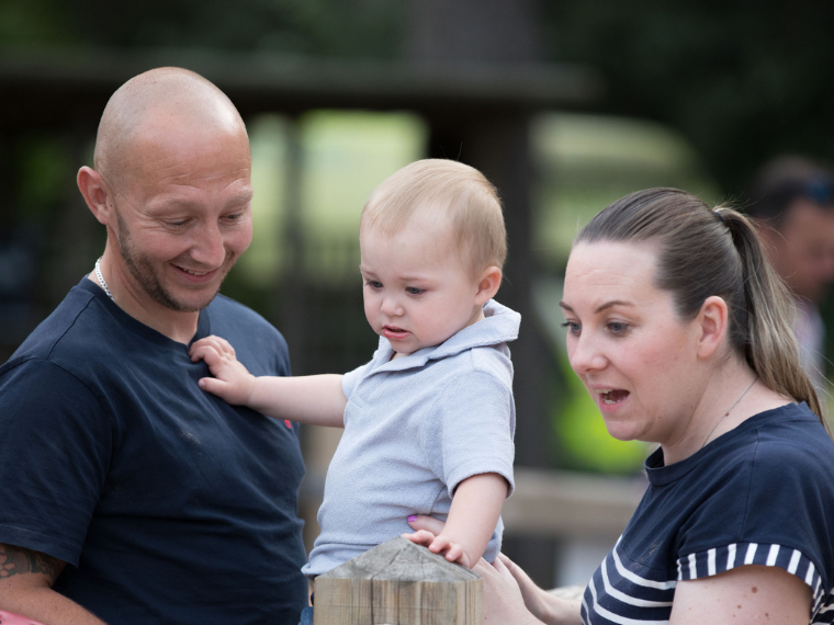 A toddler is being held by his parents. They are all looking out of shot, but they are smiling.