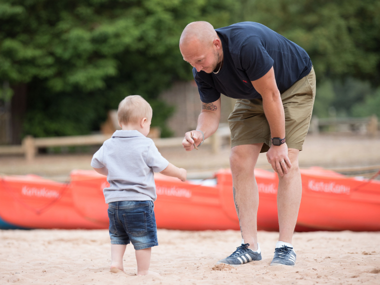 On a beach, an adult leans over to speak to the toddler in front of him